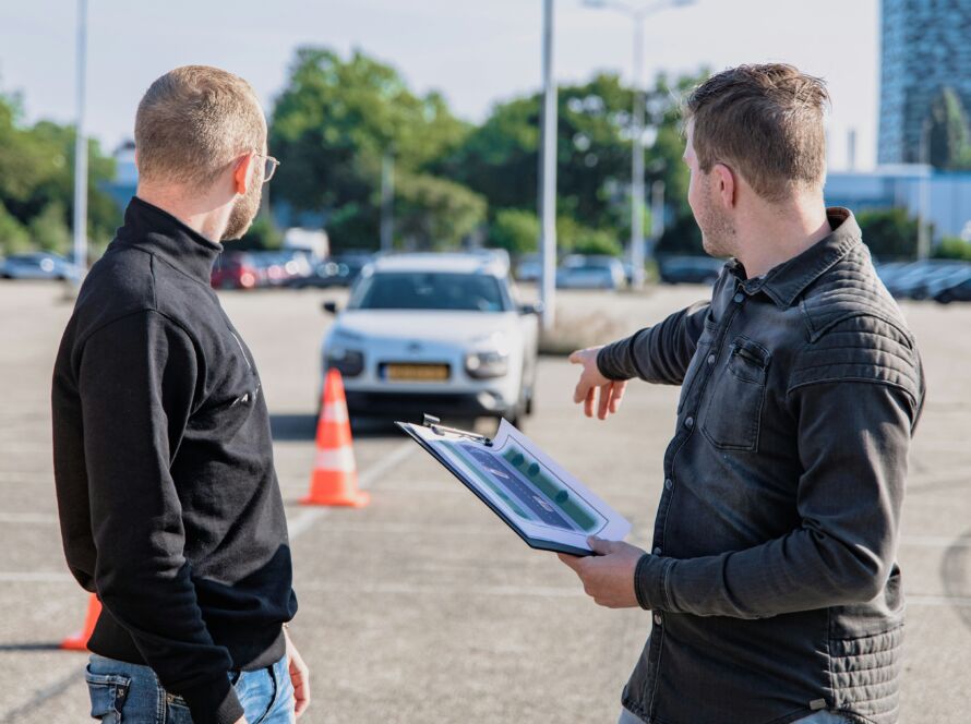 Driving instructor explaining road rules using a clipboard to a student in a parking lot, with a training car in the background