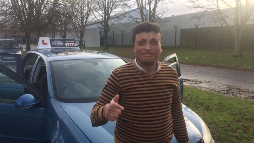 Yasser giving a thumbs up next to a blue driving school car after passing his driving test