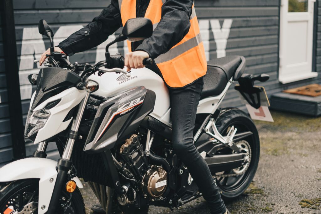 A learner motorcycle rider in a bright orange safety vest, sitting on a bike during CBT training.